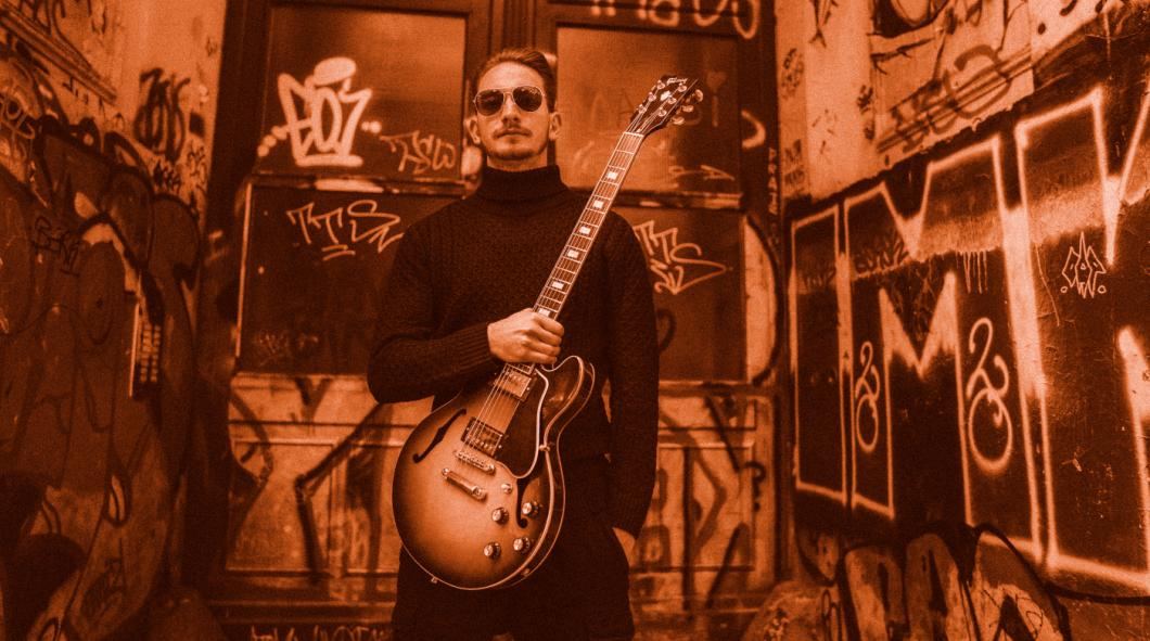 Jure Slapar with his sunburst guitar in front of a graffiti-covered doorway, in orange tones