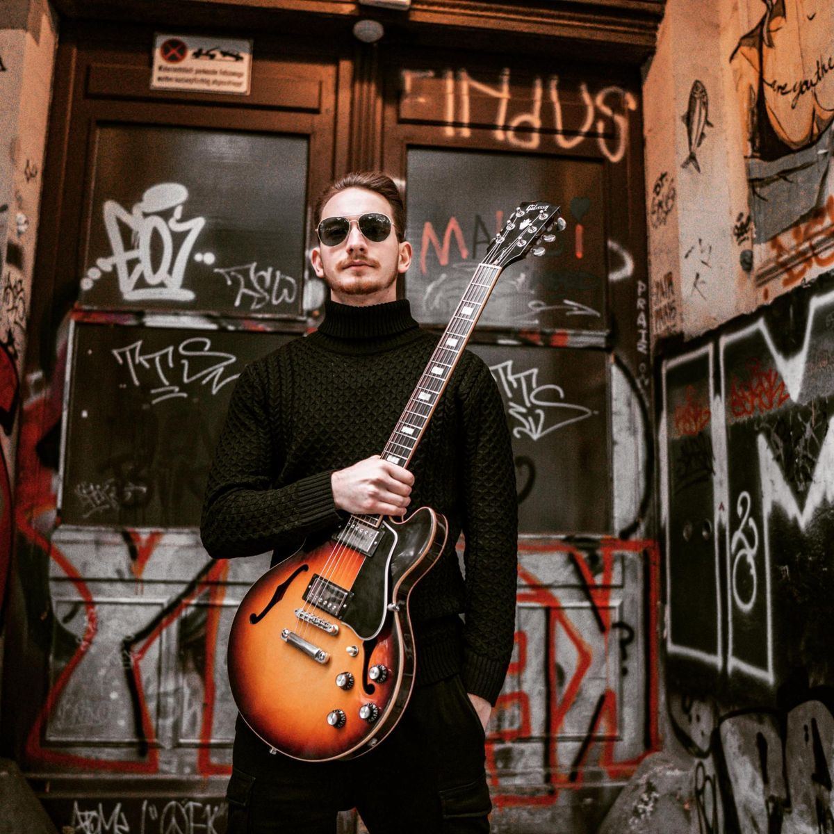 Jure Slapar standing with his sunburst Gibson semi-hollow guitar in front of a graffiti-covered doorway