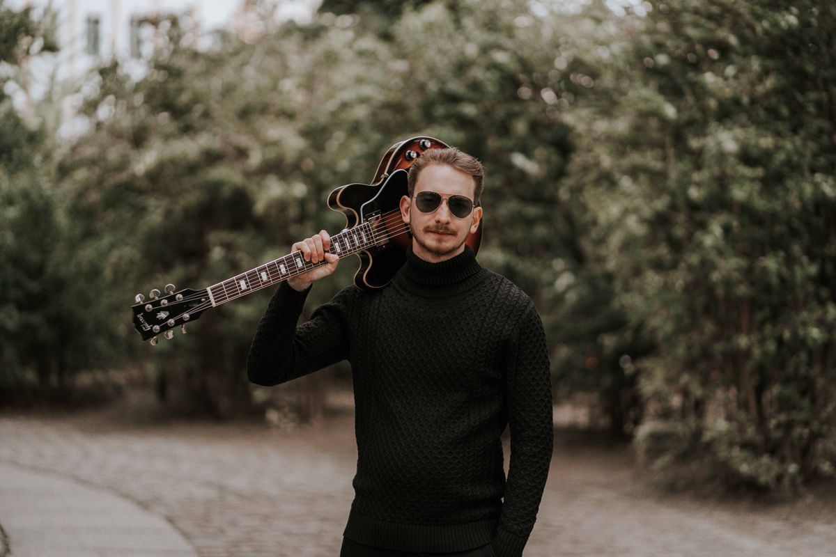 Jure Slapar with his guitar over his shoulder on a tree-lined path